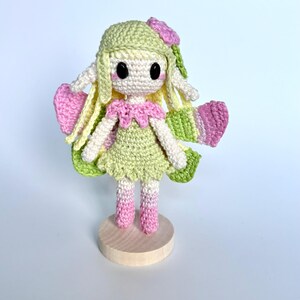May include: A crocheted doll featuring a light green dress, pink collar, and matching pink boots. The doll has long yellow hair, a light green hat with a pink flower, large black eyes, and pink cheeks. It has pink and green wings and stands on a round wooden base.