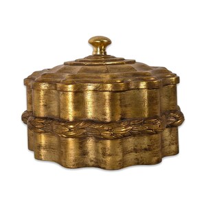 Può includere: Una scatola decorativa color oro con coperchio e pomello. La scatola ha un bordo smerlato e una superficie strutturata. Una ghirlanda dorata circonda la scatola. La scatola viene probabilmente utilizzata per riporre piccoli oggetti.
