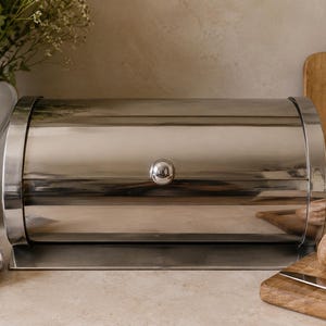 Puede incluir: Una panera de acero inoxidable pulido con tapa cilíndrica enrollable y un pequeño pomo redondo. El recipiente presenta un acabado metálico liso y reflectante con una base resistente, diseñado para almacenar pan y productos horneados.