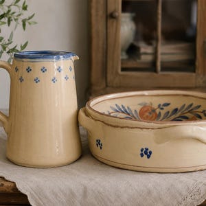May include: A cream-coloured ceramic pitcher and a matching round bowl with two handles. Both pieces feature small blue floral patterns. The pitcher has a blue rim, and the interior of the bowl displays a painted orange fruit with blue leaves.