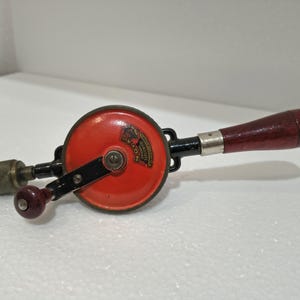 Könnte beinhalten: Eine Vintage-Handbohrmaschine mit rotem kreisförmigen Gehäuse, schwarzem Griff und Holzgriff. Der Bohrer ist messingfarben. Das Werkzeug trägt den Text "NIXON" auf dem roten Gehäuse. Ein klassisches Werkzeug für die Holzbearbeitung.