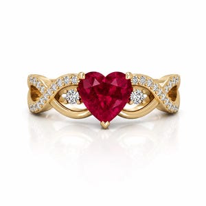Könnte beinhalten: Ein goldener Ring mit einem herzförmigen, tiefroten Edelstein als Mittelpunkt. Das Ringband hat ein Unendlichkeitsdesign, das mit kleinen, funkelnden Diamanten besetzt ist. Zwei kleinere weiße Edelsteine flankieren den herzförmigen Rubin.