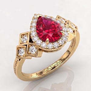 May include: A gold ring featuring a pear-shaped, deep red gemstone surrounded by a halo of smaller white stones. The ring has a decorative band with square accents and the inscription "AureviaByVarsha".