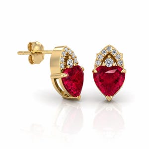 May include: A pair of gold stud earrings featuring heart-shaped red gemstones. Each earring is accented with small, clear gemstones and a decorative gold setting. The earrings are displayed on a white background.