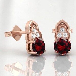 May include: A pair of rose gold stud earrings, each featuring a large round-cut deep red garnet gemstone at the base, topped with a decorative arch containing three small, clear, round-cut white diamonds set in a triangular formation.