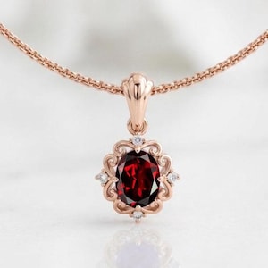 May include: A rose gold necklace featuring an oval-cut deep red garnet gemstone pendant. The pendant is surrounded by an ornate, swirling metal frame accented with four small, clear round-cut stones at the top, bottom, and sides.