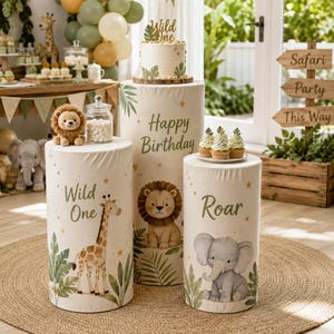 May include: Three cylindrical display pedestals featuring safari animal illustrations including a giraffe, lion, and elephant. The pedestals are printed with the text Wild One, Happy Birthday, and Roar, topped with a cake and cupcakes.