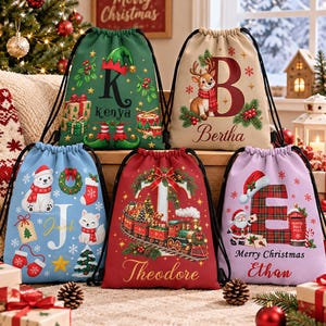 May include: Five drawstring gift bags featuring Christmas themes. The green bag has a letter K, the beige bag a letter B with a reindeer, the blue bag a letter J with a polar bear, the red bag a letter T with a train, and the lavender bag a letter E.