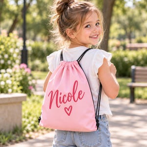 May include: A light pink drawstring backpack featuring the name Nicole in a dark pink cursive script font with a small heart icon underneath. The bag has black cord straps and is worn over the shoulders with a white short-sleeved shirt and blue jeans.