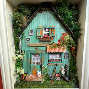 Puede incluir: Una casa de té en miniatura de color turquesa en un marco blanco, adornada con musgo, flores y pequeños accesorios. Cuenta con un toldo rojo, una pequeña puerta y una ventana con un alféizar. Incluye un pequeño letrero y una escoba en miniatura.