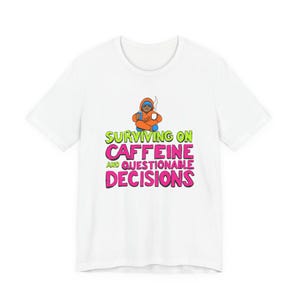 Op de afbeelding: Wit T-shirt met de tekst "SURVIVING ON CAFFEINE AND QUESTIONABLE DECISIONS" in groen en roze. De afbeelding bevat een cartoonfiguur in oranje, die een koffiekopje vasthoudt. De figuur zit in kleermakerszit.
