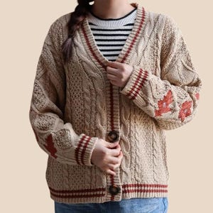 Può includere: Un cardigan beige in maglia a trecce con scollo a V, bottoni marrone scuro e bordi a righe rosse su colletto, polsini e orlo. Le maniche sono decorate con motivi ricamati di foglie d'acero rosse, indossato sopra una maglia a righe bianche e nere.
