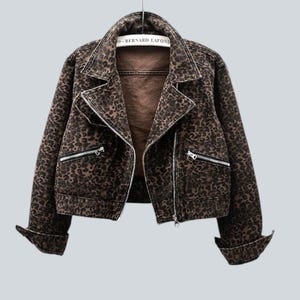 Puede incluir: Chaqueta con estampado de leopardo marrón y negro con cierre de cremallera y herrajes plateados. La chaqueta tiene un estilo motero clásico con cuello con solapas y bolsillos con cremallera. Las mangas están dobladas y la chaqueta cuelga de una percha metálica.
