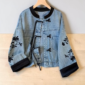 May include: Light blue denim jacket with black trim and floral embroidery on the sleeves. The jacket features a mandarin collar, frog closures, and black ties. A stylish outerwear piece.