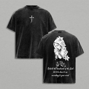 May include: A black acid-wash t-shirt featuring a small white cross graphic on the front chest. The back displays a detailed illustration of a praying figure adorned with flowers, accompanied by the text: Behold the handmaid of the Lord; let it be done to me according to your word.