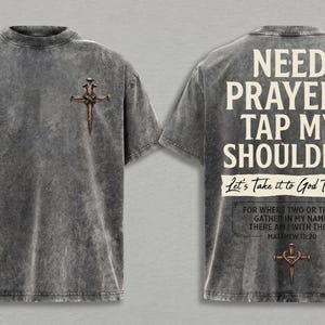 May include: A charcoal gray acid-wash t-shirt featuring a small cross graphic on the front left chest. The back displays the text NEED PRAYER? TAP MY SHOULDER. Let's Take it to God Together. FOR WHERE TWO OR THREE GATHER IN MY NAME, THERE AM I WITH THEM. MATTHEW 18:20 and a cross graphic.