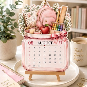May include: A decorative August 2027 calendar shaped like a mason jar, featuring a pink backpack, a red apple, a notebook, a ruler, and colourful crayons. The calendar is displayed on a small wooden easel, with a pink ribbon tied to the jar rim.