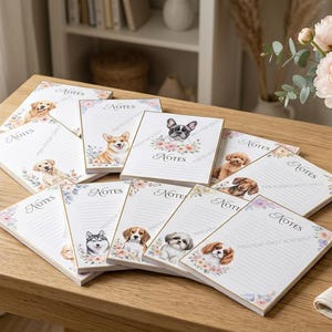 以下が含まれることがあります： 花柄のアクセントと上部に「NOTES」という文字が印刷された、様々な犬種のイラスト入り長方形メモ帳のコレクション。各メモ帳には書き込み用の横線があり、それぞれ異なる犬のポートレートが描かれています。