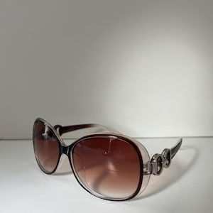 May include: Brown-tinted sunglasses with a clear frame and dark brown arms. The sunglasses have a large, oval shape and a decorative silver accent on the temples. The lenses are a gradient brown color.
