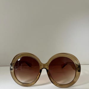 May include: Oversized round sunglasses featuring translucent brown plastic frames and gradient brown lenses. The circular lenses are encased in thick, smooth rims with a bridge connecting the two sides, creating a bold, rounded silhouette.
