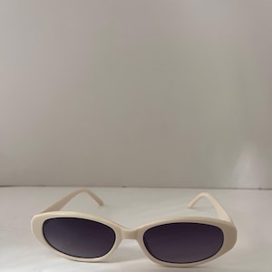 May include: Oval sunglasses featuring a smooth, cream-coloured plastic frame and dark, tinted lenses. The design includes a curved bridge and matching cream-coloured temples that extend from the sides of the frame to complete the eyewear structure.