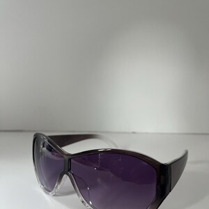 Pu&ograve; includere: Occhiali da sole viola scuro con lenti sfumate e montatura trasparente. Gli occhiali da sole hanno un design curvo e una montatura superiore marrone scuro. Le lenti sono di colore viola intenso.