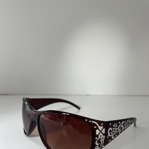 Könnte beinhalten: Braune Sonnenbrille mit einem dekorativen silbernen Blumendesign an den Bügeln. Die Gläser haben eine braune Farbverlauf. Die Sonnenbrille hat eine geschwungene, sportliche Form.