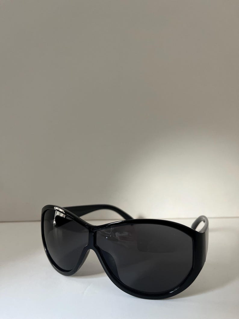 Pu&ograve; includere: Occhiali da sole neri con montatura ampia e curva e lenti scure. Gli occhiali da sole hanno un design elegante e moderno, adatto a varie forme del viso. La montatura sembra essere realizzata in un materiale lucido e resistente.