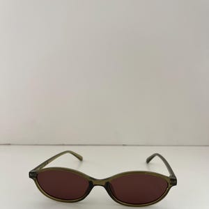 May include: Oval-shaped sunglasses with a dark olive green frame and brown lenses. The sunglasses have a sleek, minimalist design, suitable for various face shapes. The lenses provide protection from the sun's glare.