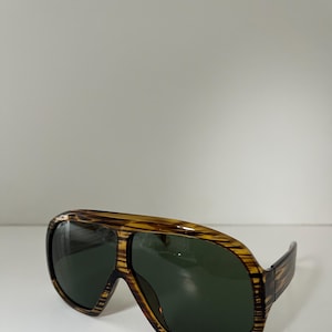 May include: Brown and yellow aviator sunglasses with dark green lenses. The frames have a striped pattern. The sunglasses are set against a white background.