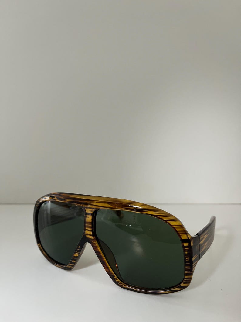 May include: Brown and yellow aviator sunglasses with dark green lenses. The frames have a striped pattern. The sunglasses are set against a white background.