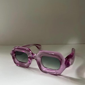 May include: A pair of stylish, translucent purple sunglasses with a unique, rounded rectangular frame. The lenses are a gradient gray, and the temples curve gracefully. The sunglasses are a fashionable accessory.