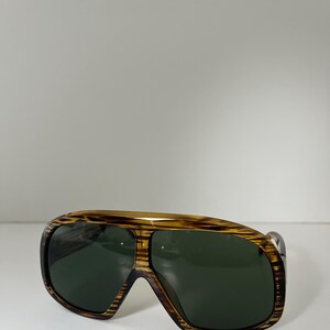 May include: Vintage sunglasses with a brown and tan striped frame and dark green lenses. The sunglasses have a wide, aviator-style shape with a thick frame.