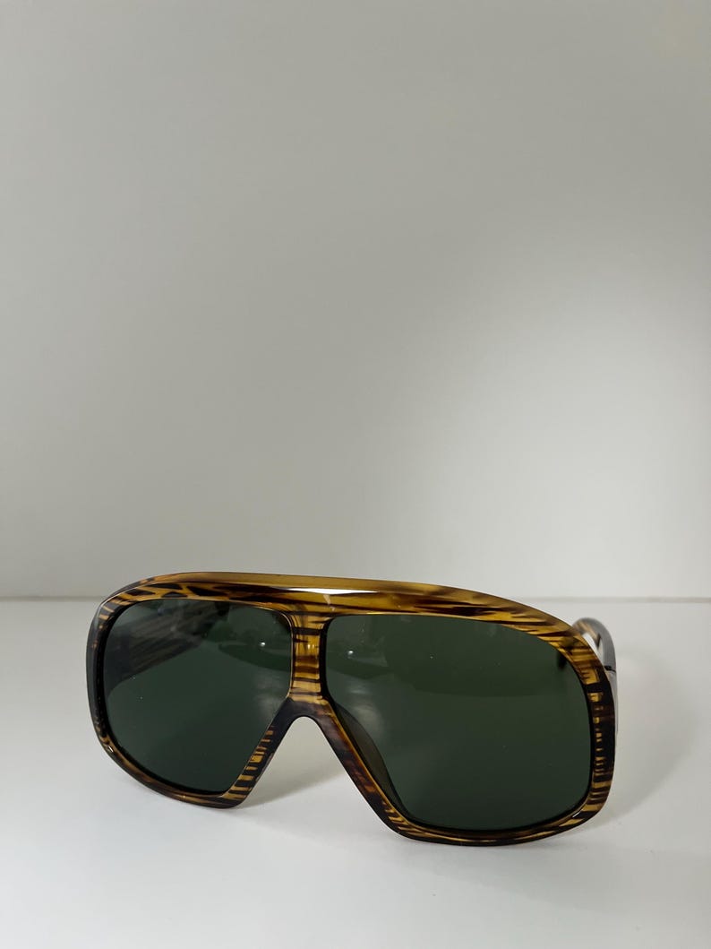 May include: Vintage sunglasses with a brown and tan striped frame and dark green lenses. The sunglasses have a wide, aviator-style shape with a thick frame.