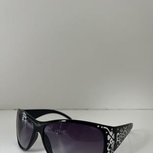 May include: Black sunglasses with a curved frame and gradient lenses. The temples feature a white floral design and small rhinestones. The sunglasses are set against a white background.
