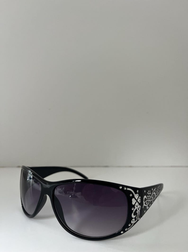 May include: Black sunglasses with a curved frame and gradient lenses. The temples feature a white floral design and small rhinestones. The sunglasses are set against a white background.