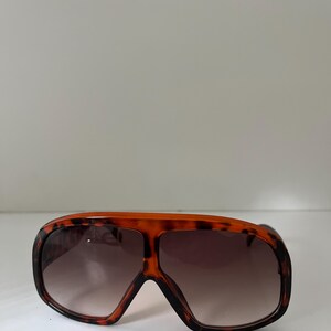 May include: Oversized sunglasses with a tortoiseshell pattern frame and gradient lenses. The sunglasses have a bold, retro design, with a thick frame and a wide, rectangular shape. The lenses transition from a dark brown at the top to a lighter shade at the bottom.