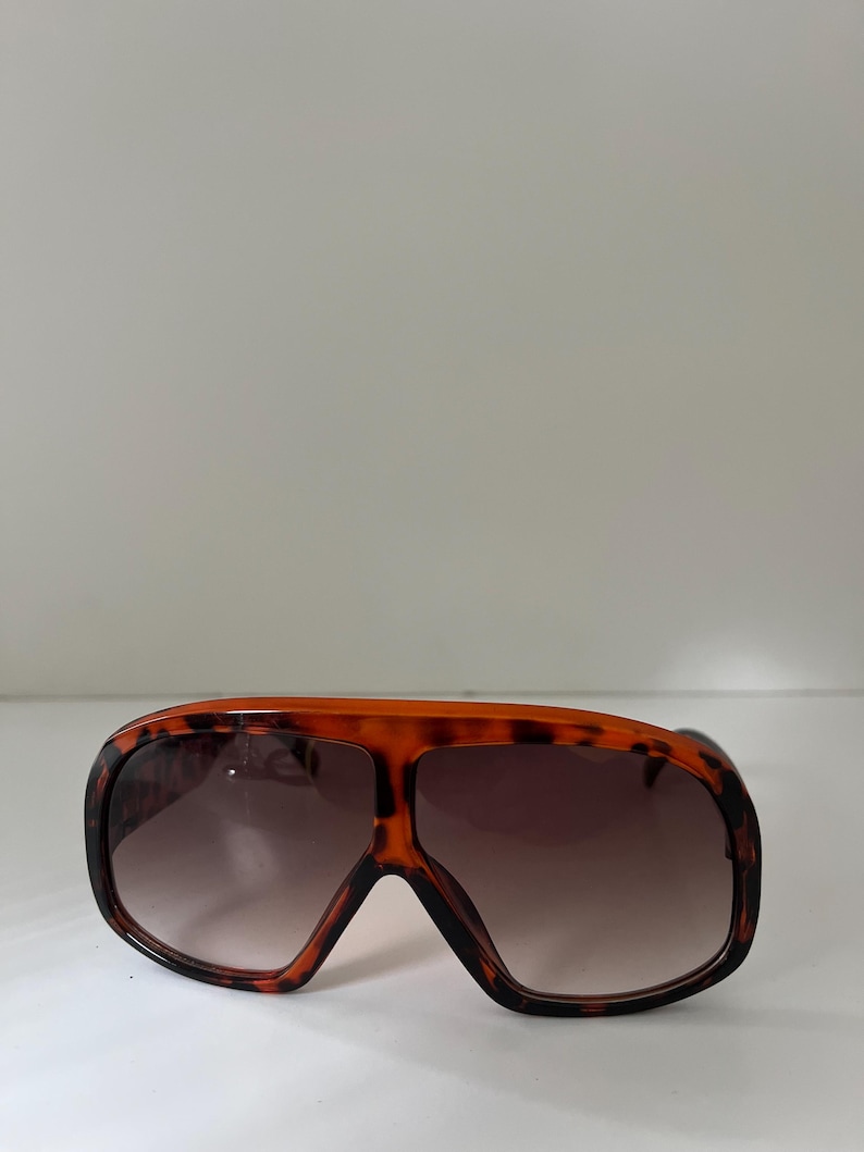 May include: Oversized sunglasses with a tortoiseshell pattern frame and gradient lenses. The sunglasses have a bold, retro design, with a thick frame and a wide, rectangular shape. The lenses transition from a dark brown at the top to a lighter shade at the bottom.
