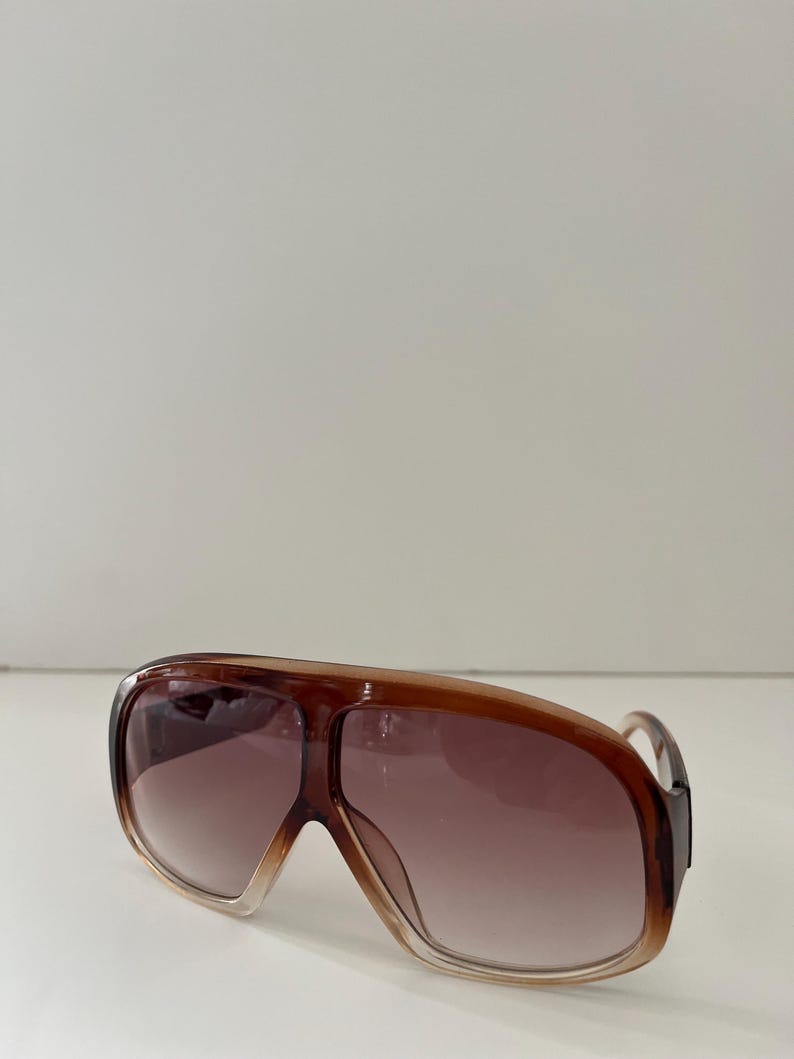May include: Brown oversized sunglasses with a gradient lens. The sunglasses have a thick frame and a classic aviator shape. The lenses transition from a light brown to a darker brown. The sunglasses are a stylish accessory.