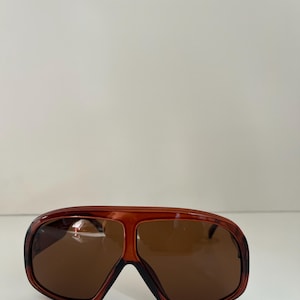 May include: Brown-tinted sunglasses with a wide, rectangular frame. The sunglasses have a dark brown frame with a lighter brown tint on the lenses. The sunglasses are a retro style.