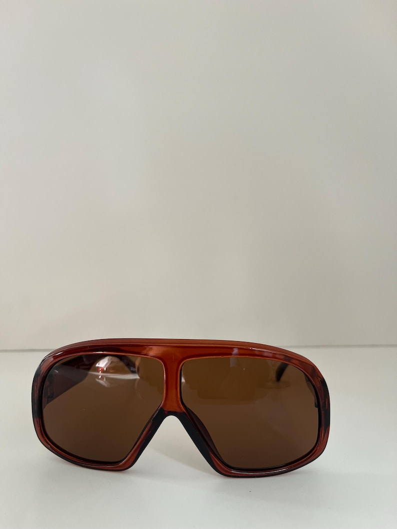 May include: Brown-tinted sunglasses with a wide, rectangular frame. The sunglasses have a dark brown frame with a lighter brown tint on the lenses. The sunglasses are a retro style.