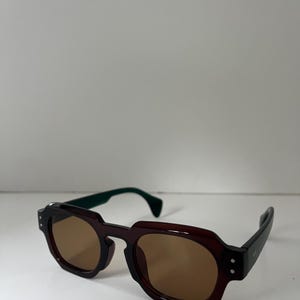 May include: Brown and black sunglasses with a geometric frame design. The sunglasses have brown lenses and green temples. The arms are black with two small silver dots on the side. The sunglasses are on a white surface.