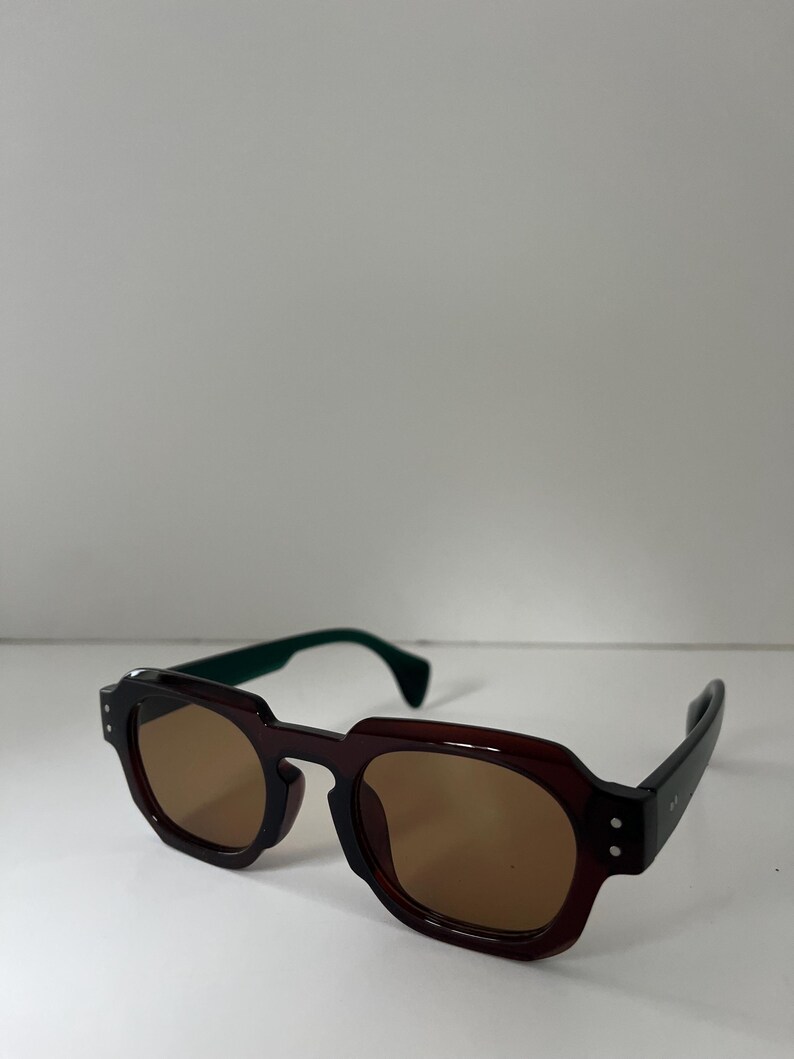 May include: Brown and black sunglasses with a geometric frame design. The sunglasses have brown lenses and green temples. The arms are black with two small silver dots on the side. The sunglasses are on a white surface.