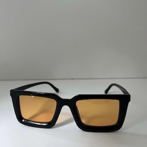 May include: Black sunglasses with a square frame and orange-tinted lenses. The sunglasses have a thick, glossy black frame and a classic, modern design. The lenses are a warm, amber color.