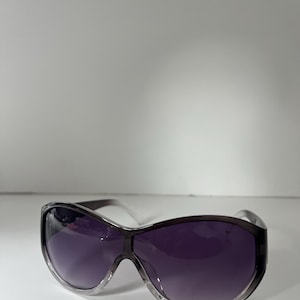 Pu&ograve; includere: Occhiali da sole oversize con lenti sfumate viola e montatura trasparente. Gli occhiali da sole hanno un design avvolgente e un ponte grigio scuro. Le lenti passano da una tonalit&agrave; pi&ugrave; chiara nella parte superiore a una tonalit&agrave; pi&ugrave; scura nella parte inferiore.