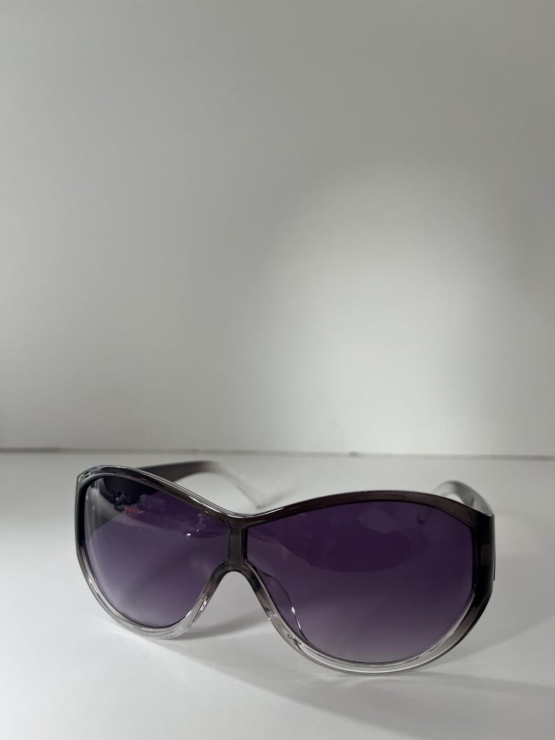 Pu&ograve; includere: Occhiali da sole oversize con lenti sfumate viola e montatura trasparente. Gli occhiali da sole hanno un design avvolgente e un ponte grigio scuro. Le lenti passano da una tonalit&agrave; pi&ugrave; chiara nella parte superiore a una tonalit&agrave; pi&ugrave; scura nella parte inferiore.