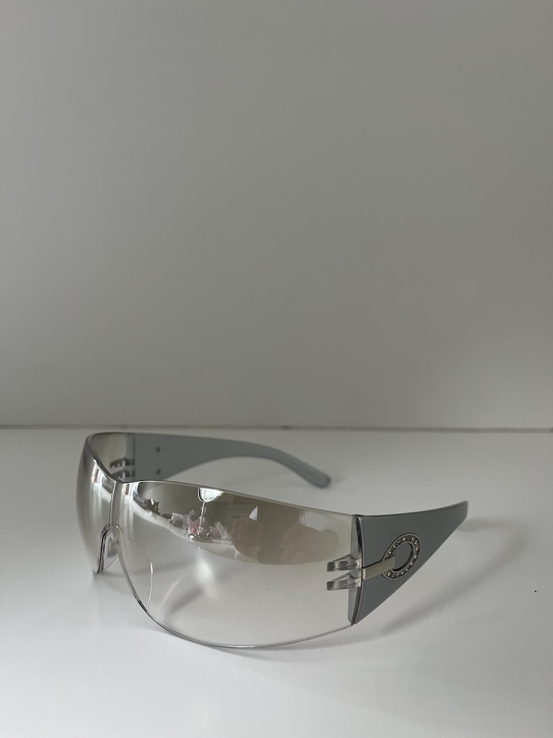 May include: Silver-framed sunglasses with a wraparound design. The lenses are clear with a slight tint, and the temples are a matching silver. The frame features a decorative element near the hinges. A modern accessory.