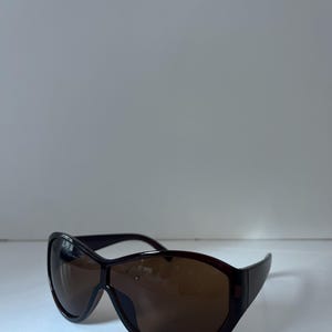Pu&ograve; includere: Occhiali da sole marrone scuro con montatura ampia e curva e lenti colorate. Gli occhiali da sole hanno un design elegante e moderno. Le lenti sono di colore marrone caldo, offrendo protezione solare.