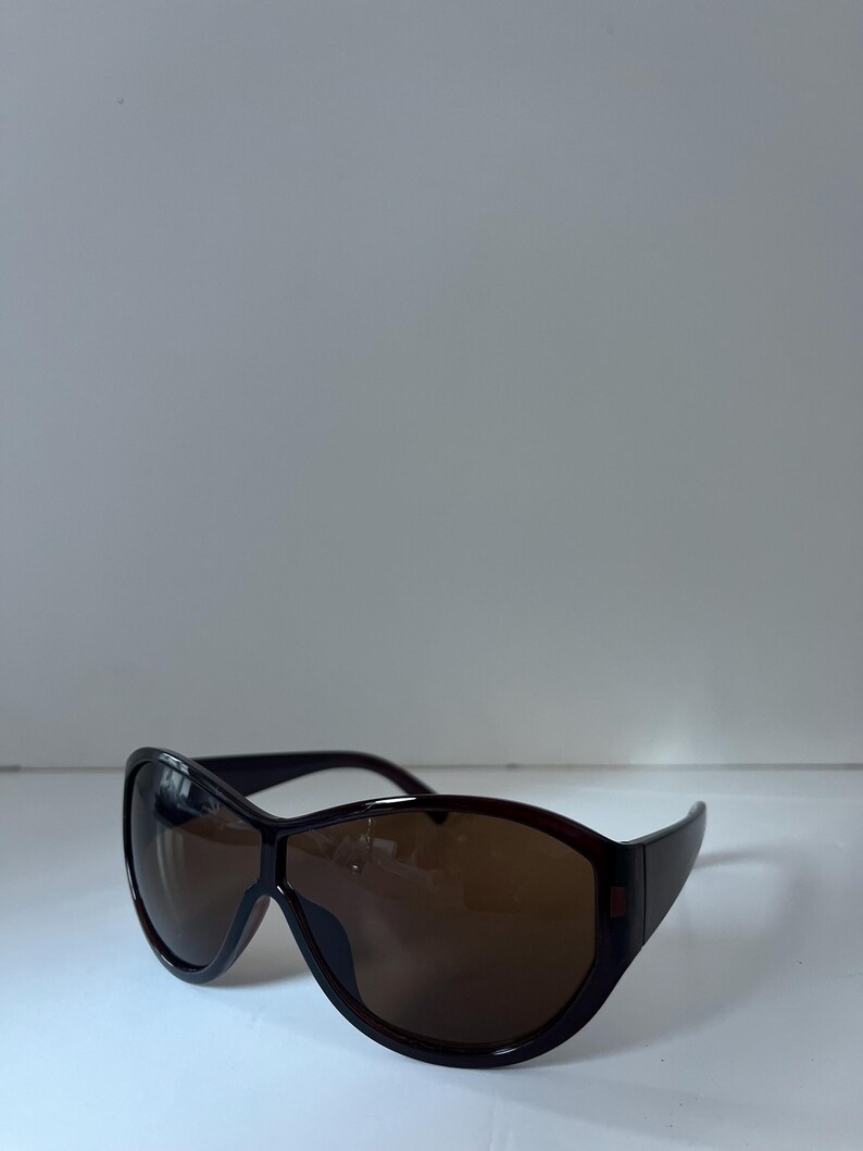Pu&ograve; includere: Occhiali da sole marrone scuro con montatura ampia e curva e lenti colorate. Gli occhiali da sole hanno un design elegante e moderno. Le lenti sono di colore marrone caldo, offrendo protezione solare.