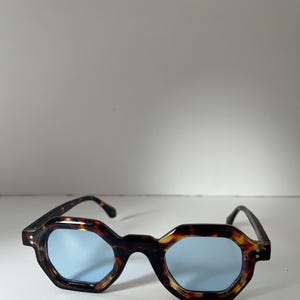 May include: Sunglasses with a tortoiseshell pattern, featuring an octagonal frame and light blue lenses. The eyewear has a dark brown and amber colour scheme, with black arms. The design offers a vintage aesthetic.